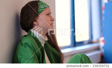 A girl in a vibrant green outfit curls into herself, her posture reflecting vulnerability and emotional contemplation. The soft lighting enhances the mood of solitude and inner reflection. 136350732