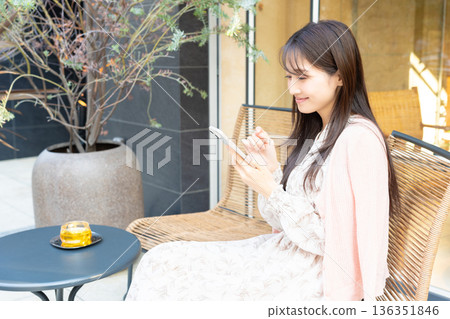 一位女士在咖啡館露台的座位上一邊喝茶一邊使用智慧型手機。 一位女士在咖啡館露台的座位上一邊喝茶一邊使用智慧型手機。 136351846