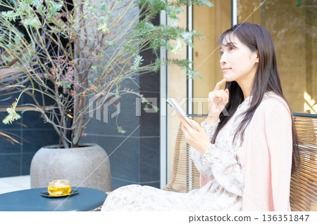 一位女士在咖啡館露台的座位上一邊喝茶一邊使用智慧型手機。 一位女士在咖啡館露台的座位上一邊喝茶一邊使用智慧型手機。 136351847