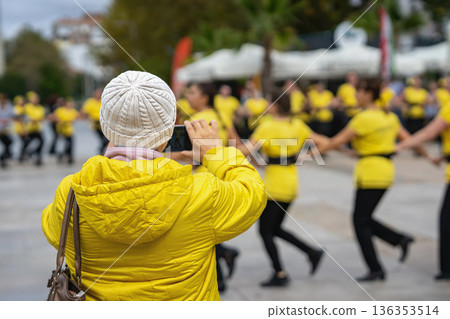Rear view of Woman in yellow jacket photographs group dance performance during outdoor public event using smartphone camera. Street culture documentation, mobile content Rear view of Woman in yellow jacket photographs group dance performance during outdoor public event using smartphone camera. Street culture documentation, mobile content 136353514
