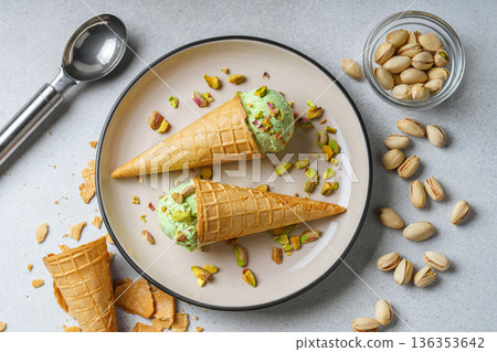 Pistachio ice cream cones on plate with nuts and scoop on gray textured background Pistachio ice cream cones on plate with nuts and scoop on gray textured background 136353642