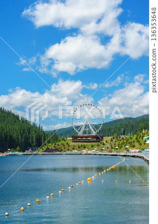 Ferris wheel near mountain lake under blue sky on summer day. Touristic resort, travel destination 136353648