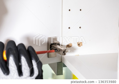 Gloved hand skillfully installs a new metal cabinet hinge into white kitchen furniture with a screwdriver, signifying essential home renovation, DIY repair, or professional assembly Gloved hand skillfully installs a new metal cabinet hinge into white kitchen furniture with a screwdriver, signifying essential home renovation, DIY repair, or professional assembly 136353980