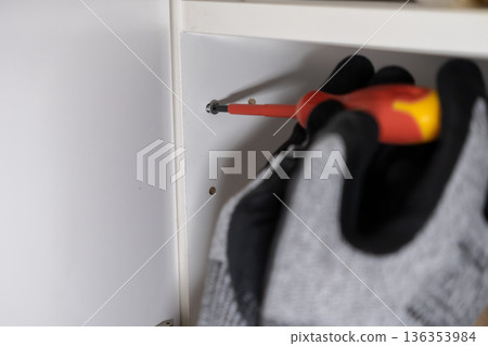 Expert gloved hand uses a red screwdriver to securely fasten a screw into sleek white furniture, symbolizing efficient home improvement, precise assembly, and DIY 136353984