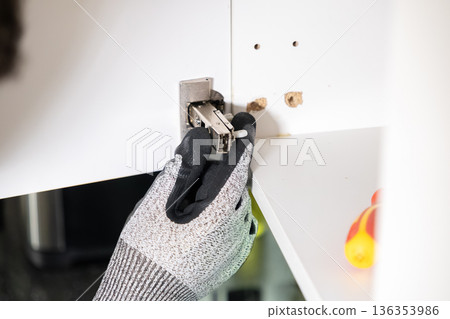 Gloved hand installing a metal cabinet hinge and plastic dowel into a white panel, representing home improvement and DIY furniture assembly 136353986