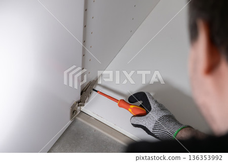 A gloved hand expertly uses a red screwdriver to install a shining metal cabinet hinge, showcasing precision and dedicated craftsmanship during home furniture assembly work 136353992