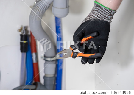 A gloved hand holds orange and black pliers near plumbing pipes, symbolizing skilled home repair and essential maintenance service for domestic systems 136353996