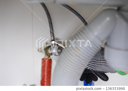Skilled plumber's gloved hand making intricate adjustments to braided water supply hoses, shut-off valve, and corrugated drain pipe connections under a kitchen sink Skilled plumber's gloved hand making intricate adjustments to braided water supply hoses, shut-off valve, and corrugated drain pipe connections under a kitchen sink 136353998