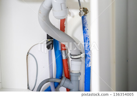 Plumbing connections with corrugated drain hoses, insulated water pipes, and cables hidden inside a cabinet, vital for domestic utility function Plumbing connections with corrugated drain hoses, insulated water pipes, and cables hidden inside a cabinet, vital for domestic utility function 136354005