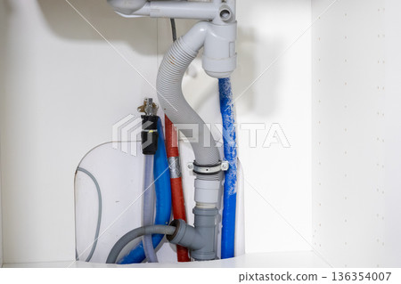 Detailed view of under sink plumbing, revealing a drain hose, red and blue water supply lines, essential for home renovation and repair projects Detailed view of under sink plumbing, revealing a drain hose, red and blue water supply lines, essential for home renovation and repair projects 136354007