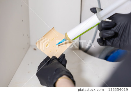 Pest control specialist in black gloves applying insecticidal gel bait to a sticky trap, managing a pest infestation in a household cabinet area, emphasizing hygiene 136354019