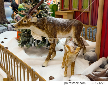 Adult and fawn reindeer figures, covered in artificial snow, stand by a twinkling Christmas tree, part of a festive holiday retail shop display 136354273