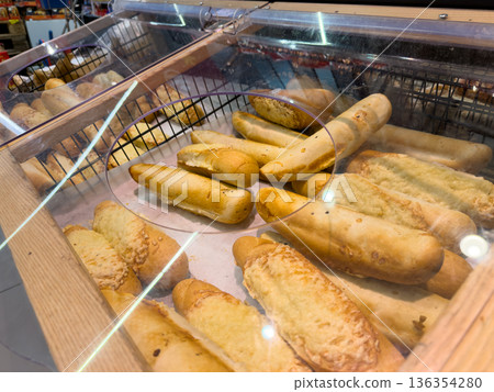 Freshly baked golden breadsticks and savory cheese-topped baguettes displayed in a modern supermarket bakery case, emphasizing daily fresh food 136354280