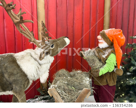 Charming reindeer statue and festive gnome figure, covered in artificial snow, stand by a hay manger, creating a magical Christmas holiday display Charming reindeer statue and festive gnome figure, covered in artificial snow, stand by a hay manger, creating a magical Christmas holiday display 136354286