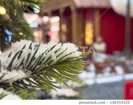 Close-up of a snow-covered evergreen branch with blurred festive lights and market stalls in the background, creating a magical winter holiday atmosphere Close-up of a snow-covered evergreen branch with blurred festive lights and market stalls in the background, creating a magical winter holiday atmosphere 136354320