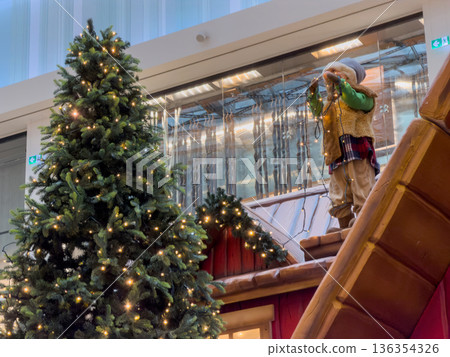 A festive Christmas tree brightly lit with warm lights stands next to a charming decorative doll on a miniature rooftop, creating a joyful holiday display A festive Christmas tree brightly lit with warm lights stands next to a charming decorative doll on a miniature rooftop, creating a joyful holiday display 136354326