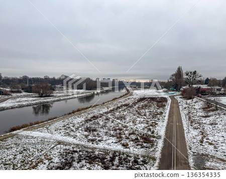Overcast winter landscape featuring a tranquil calm river, snow-dusted banks, and a winding pathway leading toward a distant city under a grey sky, highlighting urban nature Overcast winter landscape featuring a tranquil calm river, snow-dusted banks, and a winding pathway leading toward a distant city under a grey sky, highlighting urban nature 136354335