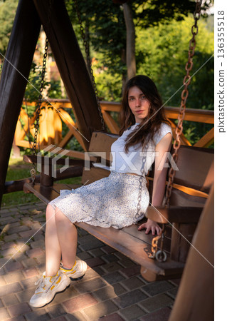 Young woman relaxing on wooden swing bench in garden at sunset Young woman relaxing on wooden swing bench in garden at sunset 136355518