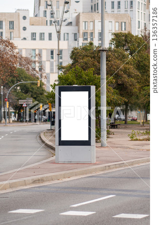 Blank digital signage billboard presenting a white screen for advertising, located on a busy city street by a road with buildings and trees in the background 136357186