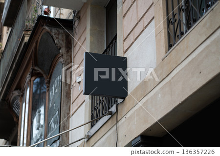 Blank dark mockup sign projecting from an old European building facade with decorative wrought iron balcony railing, providing clear branding opportunity in urban street architecture 136357226