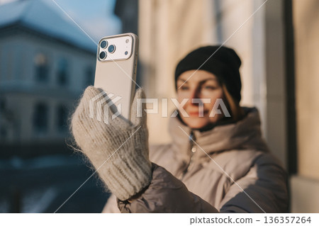 Woman takes selfie outside in winter with a mobile phone while wearing a warm jacket and hat 136357264