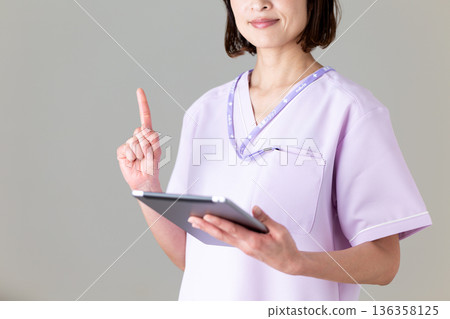 A middle-aged female nurse wearing a medical scrub holding a tablet and pointing her index finger up 136358125