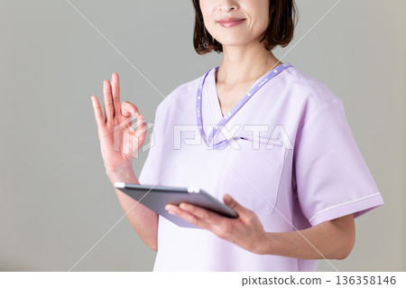 A middle-aged female nurse wearing a medical scrub holding a tablet and making a circle sign 136358146