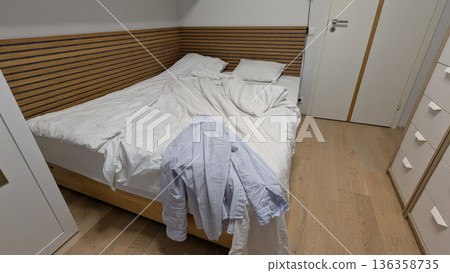 Unmade bed with white bedding in cloud dancer color of the year 2026, blue striped shirt on corner of wooden bed, cozy real bedroom interior with natural wood floor and soft lighting 136358735