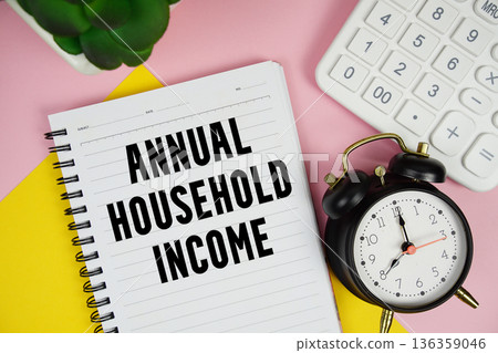 Annual Household Income text written on notepad with calculator and alarm clock on pink background 136359046