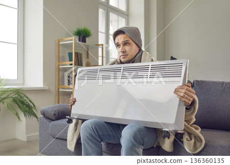 Frozen man sitting on sofa wrapped in a blanket at home and holding electric heater in hands. 136360135