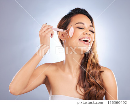 Happy woman, face roller and massage for skincare, rose quartz cosmetics or salon dermatology on studio background. Female model, crystal stone tools or smile for facial, beauty or lymphatic drainage Happy woman, face roller and massage for skincare, rose quartz cosmetics or salon dermatology on studio background. Female model, crystal stone tools or smile for facial, beauty or lymphatic drainage 136360492