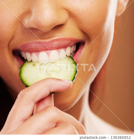 Woman, mouth or eating a cucumber for skincare nutrition or healthy diet against a brown studio background. Smile, bite or closeup of girl vegetable for organic facial treatment or natural cosmetics Woman, mouth or eating a cucumber for skincare nutrition or healthy diet against a brown studio background. Smile, bite or closeup of girl vegetable for organic facial treatment or natural cosmetics 136360852