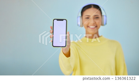Happy woman with phone screen, headphones and mockup in studio for social media post, mobile app and streaming radio. Smile, music media subscription and girl with cellphone on blue background space. Happy woman with phone screen, headphones and mockup in studio for social media post, mobile app and streaming radio. Smile, music media subscription and girl with cellphone on blue background space. 136361322