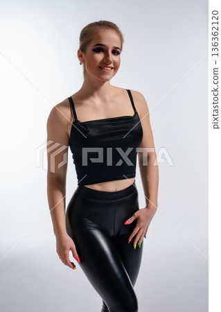 Stylish young woman in black faux leather leggings and crop top posing confidently in a studio. 136362120
