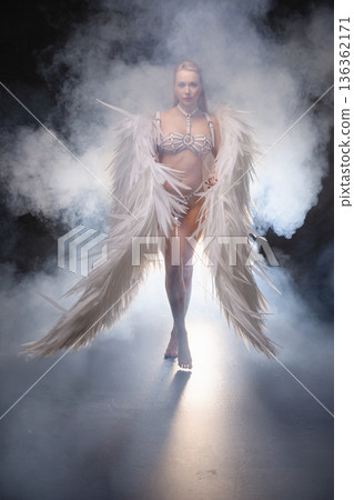 Powerful woman in white angel costume with large feather wings walking through dramatic smoke. Powerful woman in white angel costume with large feather wings walking through dramatic smoke. 136362171
