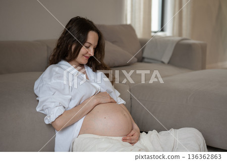 Pregnant woman caressing her belly sitting near the sofa at home 136362863