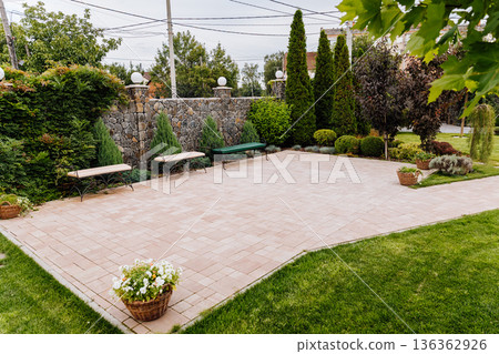 Paved patio plaza with benches in landscaped garden, stone wall, lawn and evergreen trees Paved patio plaza with benches in landscaped garden, stone wall, lawn and evergreen trees 136362926
