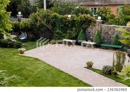 Spacious paved patio with benches in landscaped backyard, stone wall, lawn and shrubs Spacious paved patio with benches in landscaped backyard, stone wall, lawn and shrubs 136362960