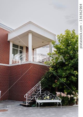 Brick house facade with balcony, white columns, wrought iron railing and garden bench Brick house facade with balcony, white columns, wrought iron railing and garden bench 136362964