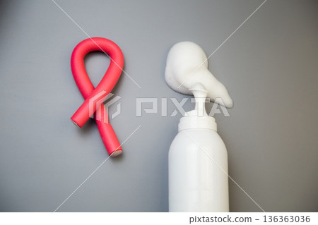 A close-up on a gray background shows a bottle of foam for long-term hair styling, white foam poured out of it, next to it lies a curved pink curler. The concept of styling, hairstyle, curls. High 136363036