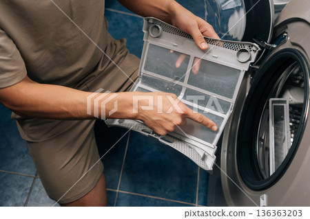 Person removing lint from tumble dryer filter, performing routine household chore and essential appliance maintenance, ensuring proper functioning and preventing fire hazards. Person removing lint from tumble dryer filter, performing routine household chore and essential appliance maintenance, ensuring proper functioning and preventing fire hazards. 136363203