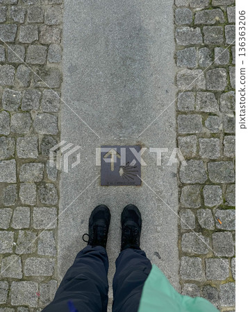 Pilgrim standing on Camino de Santiago waymark with yellow arrow and shell symbol on stone pavement. Pilgrimage, hiking journey, faith, and long-distance walking route in Europe 136363206