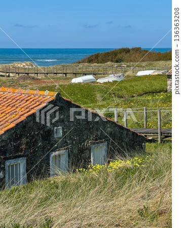 Stone coastal house with red tiled roof surrounded by dune grass and wildflowers, wooden boardwalk and Atlantic Ocean. Travel reporting, coastal geography documentation, regional architecture Stone coastal house with red tiled roof surrounded by dune grass and wildflowers, wooden boardwalk and Atlantic Ocean. Travel reporting, coastal geography documentation, regional architecture 136363208