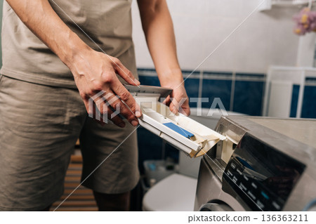 Cropped shot of man opening washing machine detergent dispenser drawer in the bathroom, pulling out the compartment to add laundry detergent for washing clothes. Cropped shot of man opening washing machine detergent dispenser drawer in the bathroom, pulling out the compartment to add laundry detergent for washing clothes. 136363211