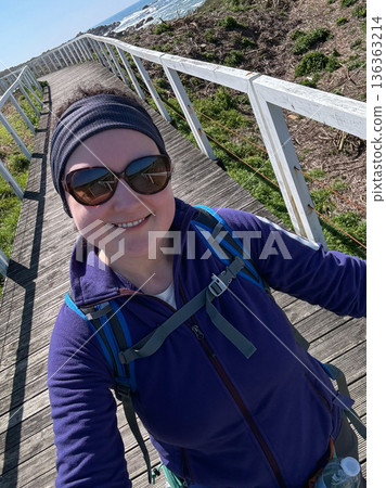 Solo female pilgrim walking boardwalk with backpack, outdoor clothing, sunglasses, capturing selfie on coastal Camino de Santiago. Travel marketing, solo travel, hiking lifestyle content, pilgrimage Solo female pilgrim walking boardwalk with backpack, outdoor clothing, sunglasses, capturing selfie on coastal Camino de Santiago. Travel marketing, solo travel, hiking lifestyle content, pilgrimage 136363214