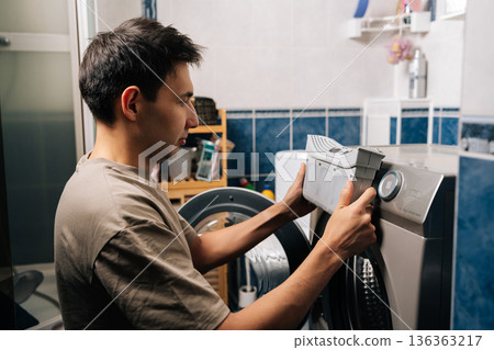 Man holding grey lint filter, removing from silver tumble dryer in laundry room, performing essential appliance cleaning and maintenance to ensure proper function and efficiency. 136363217