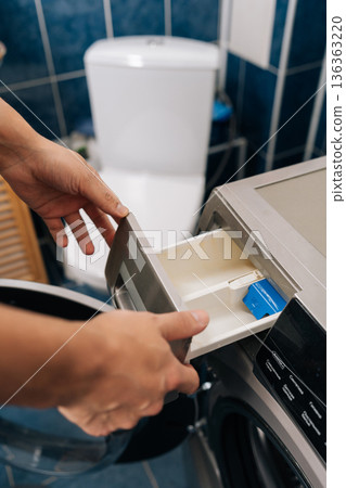 Vertical shot of hands opening empty powder detergent tray of modern front-loading washing machine in bathroom, preparing for laundry and daily household cleaning routine. Concept of household chores. 136363220