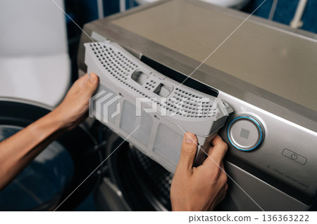 Man removing lint filter from modern tumble dryer, performing routine appliance maintenance for optimal performance and household safety, preventing fire hazards and improving laundry efficiency. 136363222