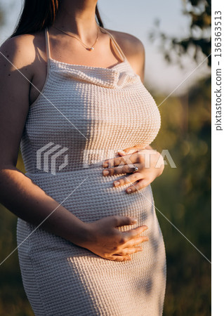 Pregnant woman in beige dress holding baby bump at sunset outdoors Pregnant woman in beige dress holding baby bump at sunset outdoors 136363513