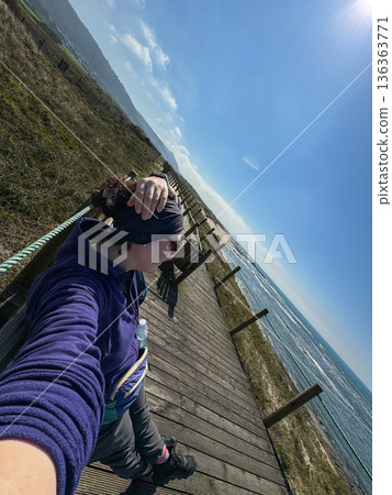 Solo female hiker walking wooden boardwalk along Atlantic coast, backpack travel on Camino de Santiago route in Portugal. Travel marketing, adventure tourism, solo travel storytelling 136363771